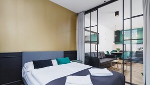 A modern hotel room with a large bed, a sofa, and a view through a glass door to a dining area.