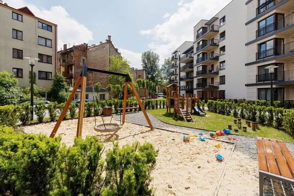 A playground with swings and slides, surrounded by neatly trimmed bushes and a sandy area.