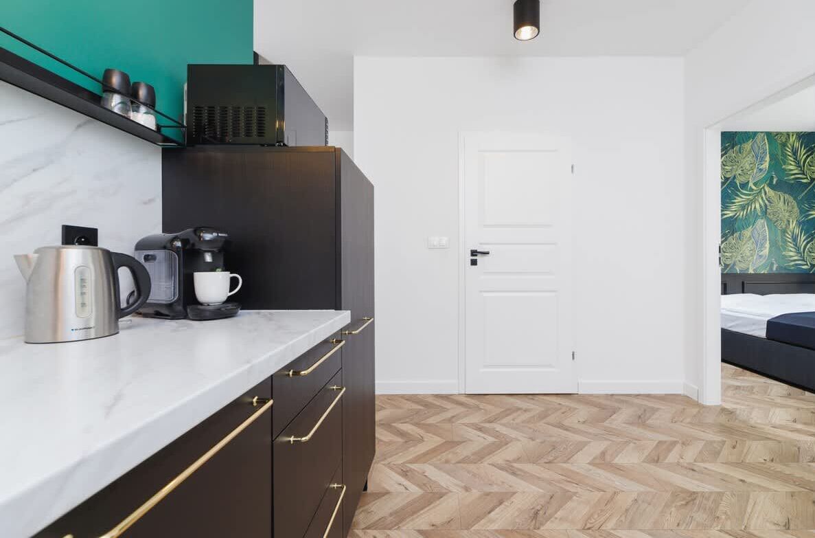 A modern kitchen with a marble countertop, a coffee maker, and a kettle. There is a white door and a bed with a green leaf-patterned wall in the background.