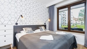 Un dormitorio moderno con pared de diseño geométrico, una cama grande con colcha gris y blanca, dos lámparas de noche, un aparador blanco y una ventana con vistas a edificios.