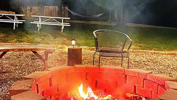 Una zona con fogones, asientos y una mesa de picnic, rodeada por un campo cubierto de césped y una estructura de madera al fondo.