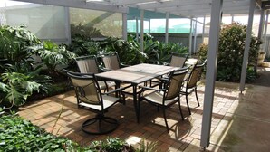Un patio cubierto con una mesa y sillas, rodeado de plantas y vistas a un edificio al fondo.