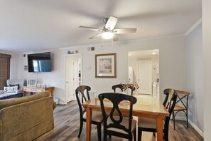 A dining area with a wooden table and chairs, a television mounted on the wall, and a ceiling fan. - 871 Ketch Court II 1BR 1BA 1ST Floor Walk-In Shower Patio Pool Gas Grill (Hilton Head Island)