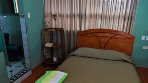 A hotel room with a bed, a small wooden side table with a telephone and a vase with a rose, a bathroom with a toilet and a shower, and a curtain with a pattern. - Hospedaje Don Juan (Cusco)