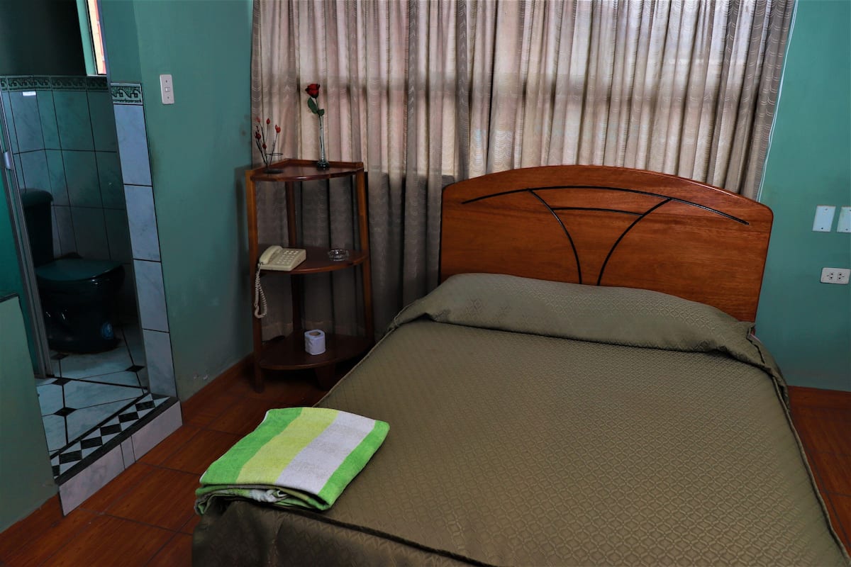 A hotel room with a bed, a small wooden side table with a telephone and a vase with a rose, a bathroom with a toilet and a shower, and a curtain with a pattern.