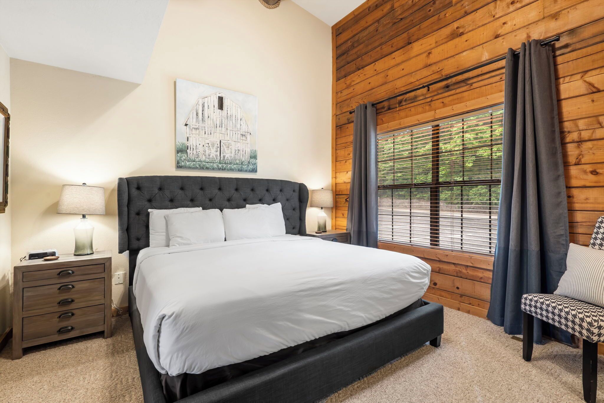 Une chambre avec un grand lit, une tête de lit en bois, une table de chevet avec une lampe, une chaise et une fenêtre avec des rideaux.