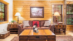A cozy living room with a wooden wall, a framed picture of a bear, a brown leather sofa, two lamps, and a wooden coffee table. - Lovely Log Cabin with Whirlpool Tub in Branson Theatre District (Branson)