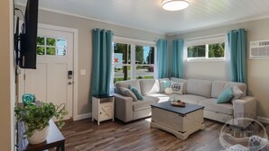 A spacious living room with a beige sofa, a coffee table, and a TV mounted on the wall. There are windows with curtains, a door, and a small side table with a plant. - Torch Lake Sandbar Directly Across the Street With Indoor Boat Slip Unit A (Rapid City)