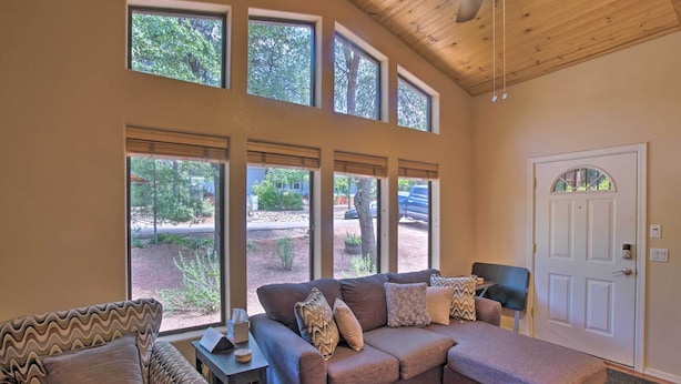 A living room with a brown sofa, a patterned armchair, and a small table with a lamp. There are large windows with blinds, a door, and a ceiling fan. - Forest-View Retreat in Payson, Close to Hiking! (Payson)