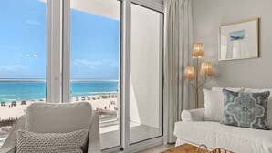 Una habitación con un amplio ventanal corredizo hacia la playa, un sofá blanco, una silla y una mesa de centro con una revista.