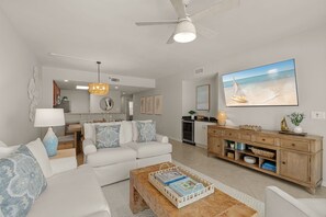 A living room with a white sofa, a coffee table, a wooden cabinet, and a large TV displaying a beach scene. - Downtown Destin Condo on the Beach (Destin)