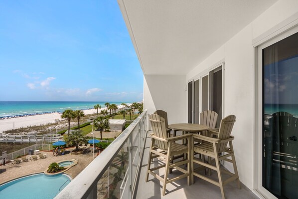 A balcony with a table and chairs, overlooking a beach and a pool.
