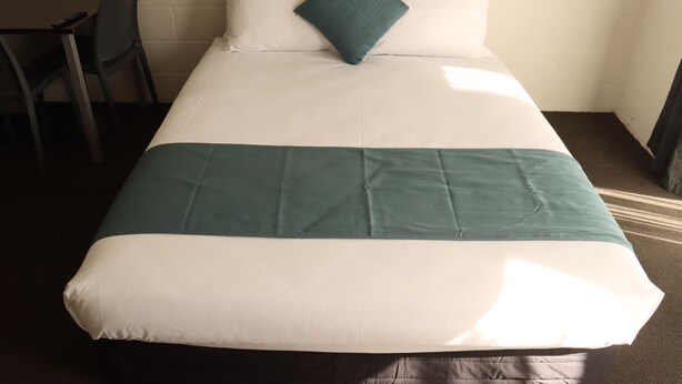 Un letto rifatto con una coperta bianca e color petrolio, un cuscino color petrolio e una sedia alla sinistra.