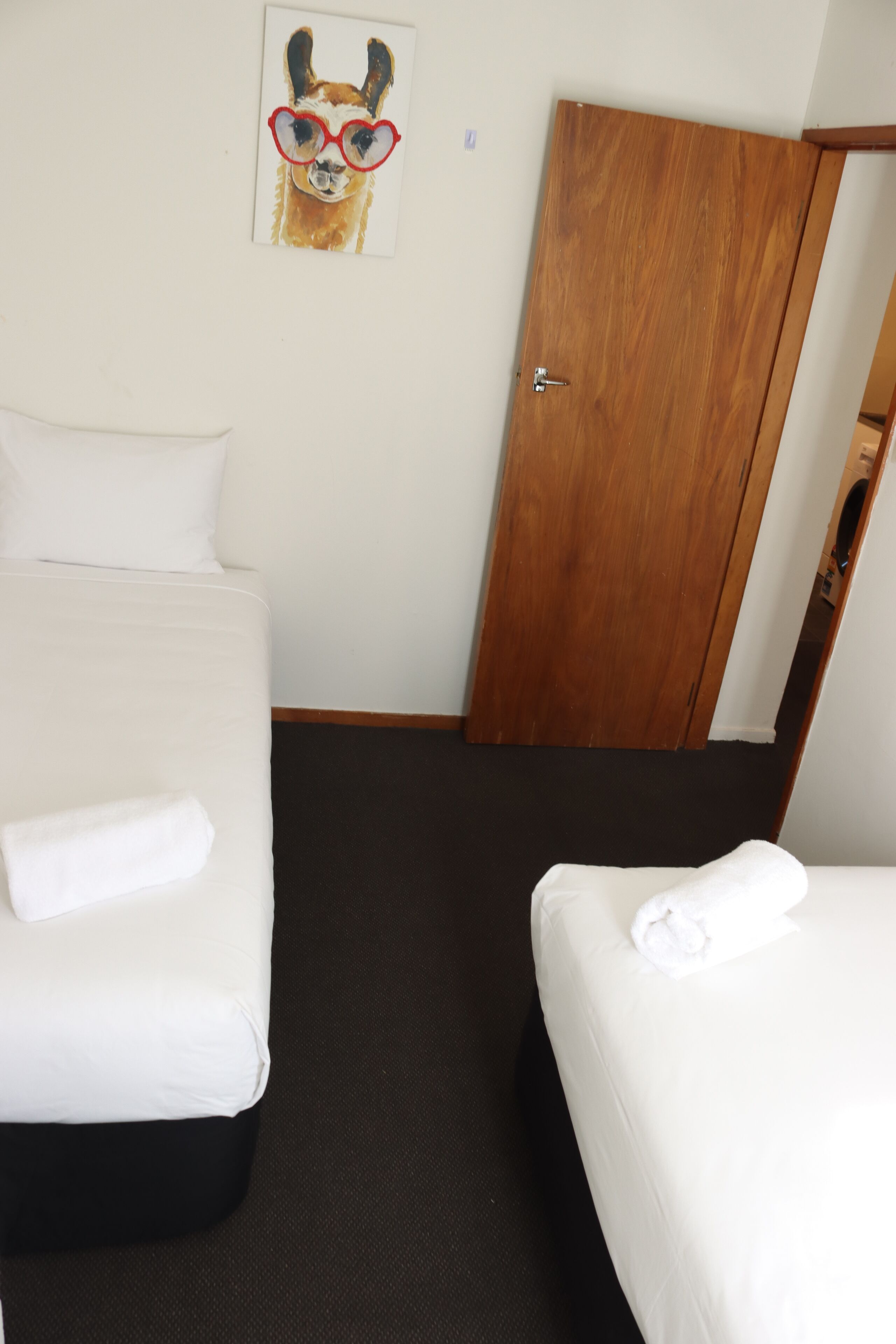 Una habitación de hotel con dos camas, una puerta de madera y un cuadro de un perro con gafas.