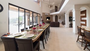 Une salle à manger spacieuse avec une longue table dressée pour le repas, un plafond haut avec un ventilateur et une vue sur l’extérieur.