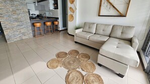 A modern living room with a beige sofa, a kitchen in the background, and a decorative wall hanging. - VistaPinero Beach Front Apartment - Joyudas Cabo Rojo (Cabo Rojo)