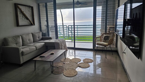 A modern living room with a grey sofa, a coffee table, and a wicker chair. Large windows offer a view of a balcony with a railing. - VistaPinero Beach Front Apartment - Joyudas Cabo Rojo (Cabo Rojo)