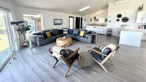 Un salon moderne avec un canapé d’angle, un pouf rond et une table basse. On aperçoit également un coin repas avec une cuisine en arrière-plan.