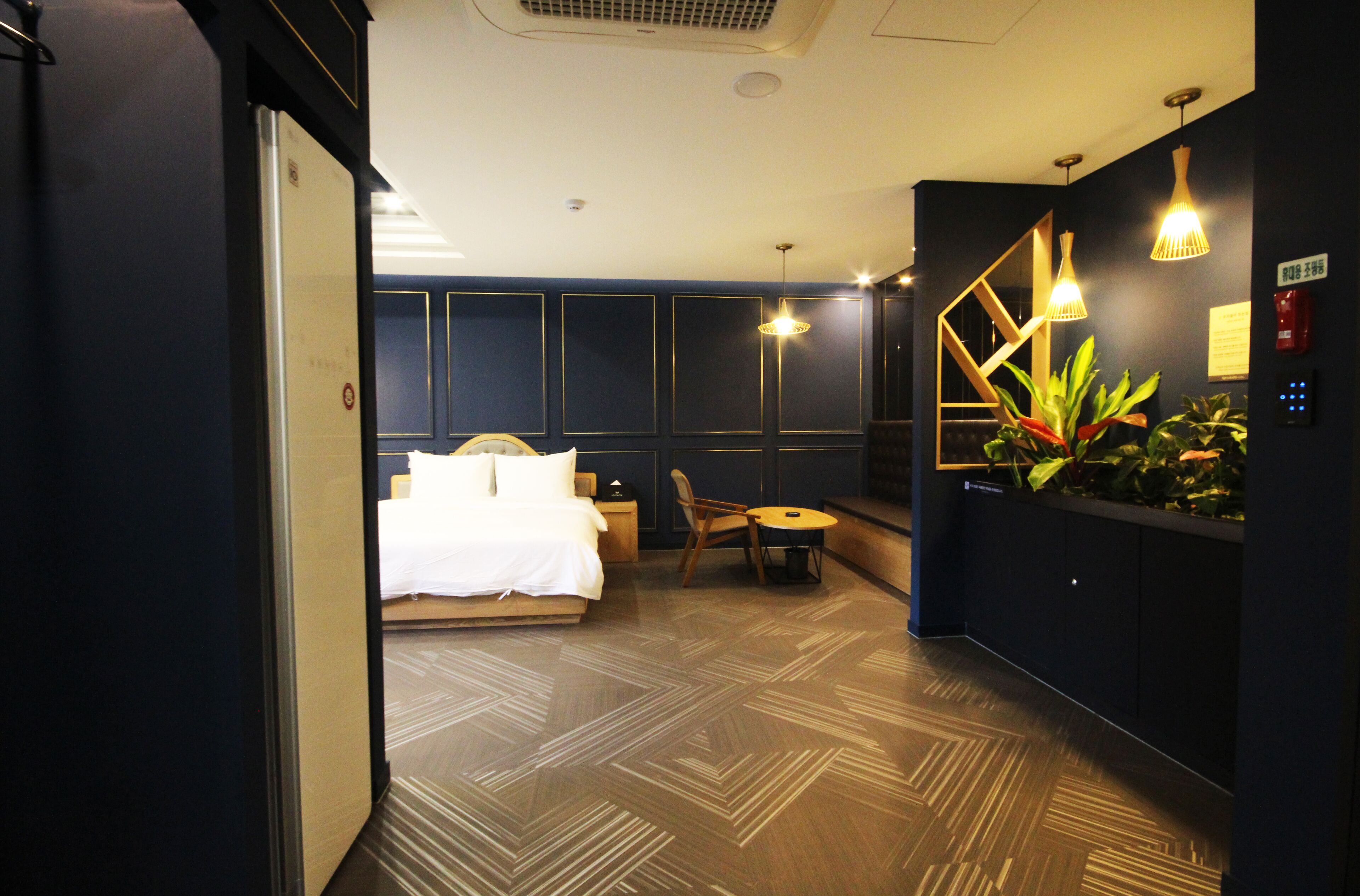 Habitación de hotel con cama, mesita de noche, silla y una pequeña mesa. Hay dos lámparas colgantes y una planta en maceta en la pared.