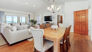 A spacious living area with a wooden dining table, white sofa, and a large TV.