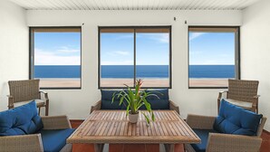 A room with a wooden table and wicker chairs, a potted plant, and large windows with a beach view.
