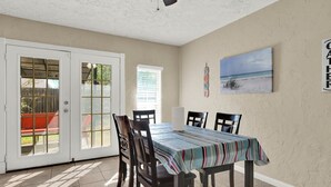 Uma sala de jantar com uma mesa posta para quatro pessoas, rodeada por cadeiras, um ventilador de teto e uma pintura de uma paisagem praiana.