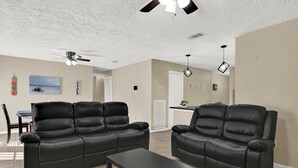 Uma sala de estar com dois sofás de couro preto, uma mesa de centro, um ventilador de teto e uma cozinha ao fundo.