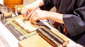 En person som tillagar sushi i ett kök med en skärbräda i trä, en skål i metall och en behållare i metall.