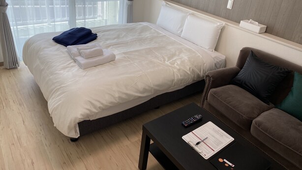 A hotel room with a large bed, a brown sofa, a black coffee table with a clipboard and pen, and a window with curtains. - HOTEL Ishigakijima 2020 (Ishigaki)