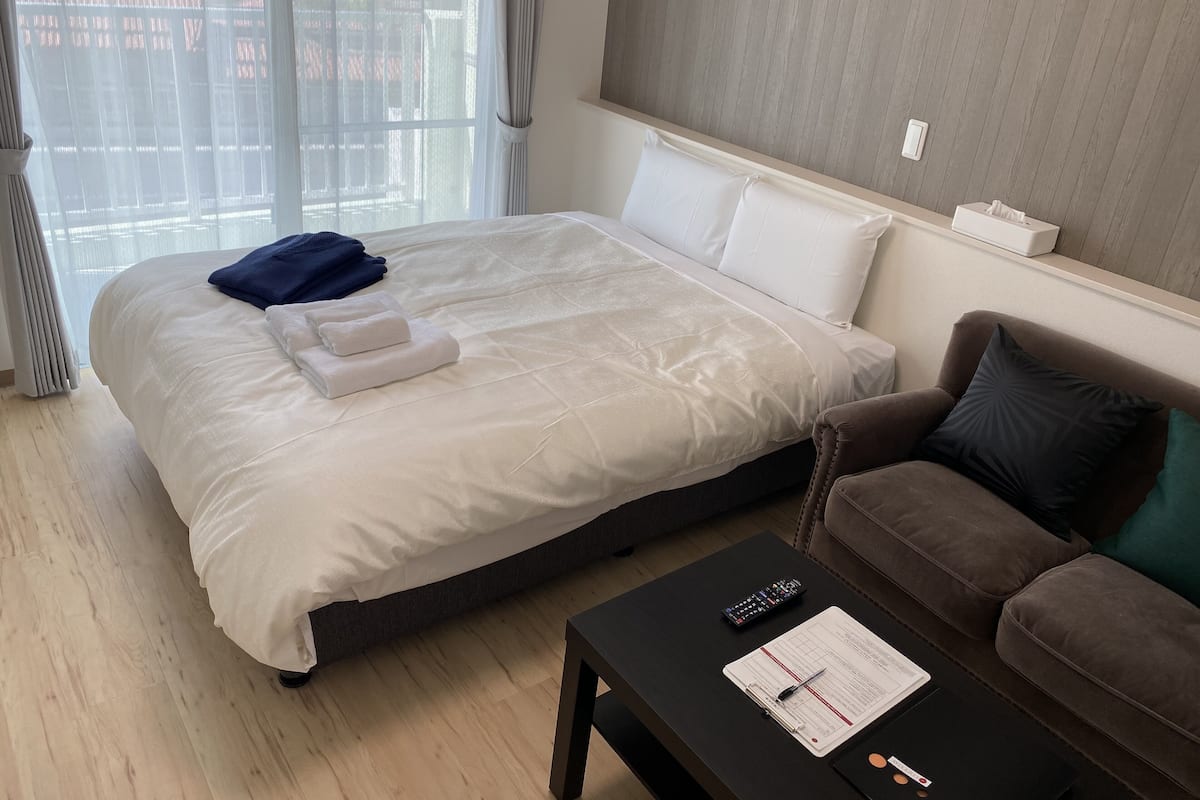 A hotel room with a large bed, a brown sofa, a black coffee table with a clipboard and pen, and a window with curtains.