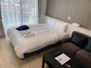 A hotel room with a large bed, a brown sofa, a black coffee table with a clipboard and pen, and a window with curtains. - HOTEL Ishigakijima 2020 (Ishigaki)