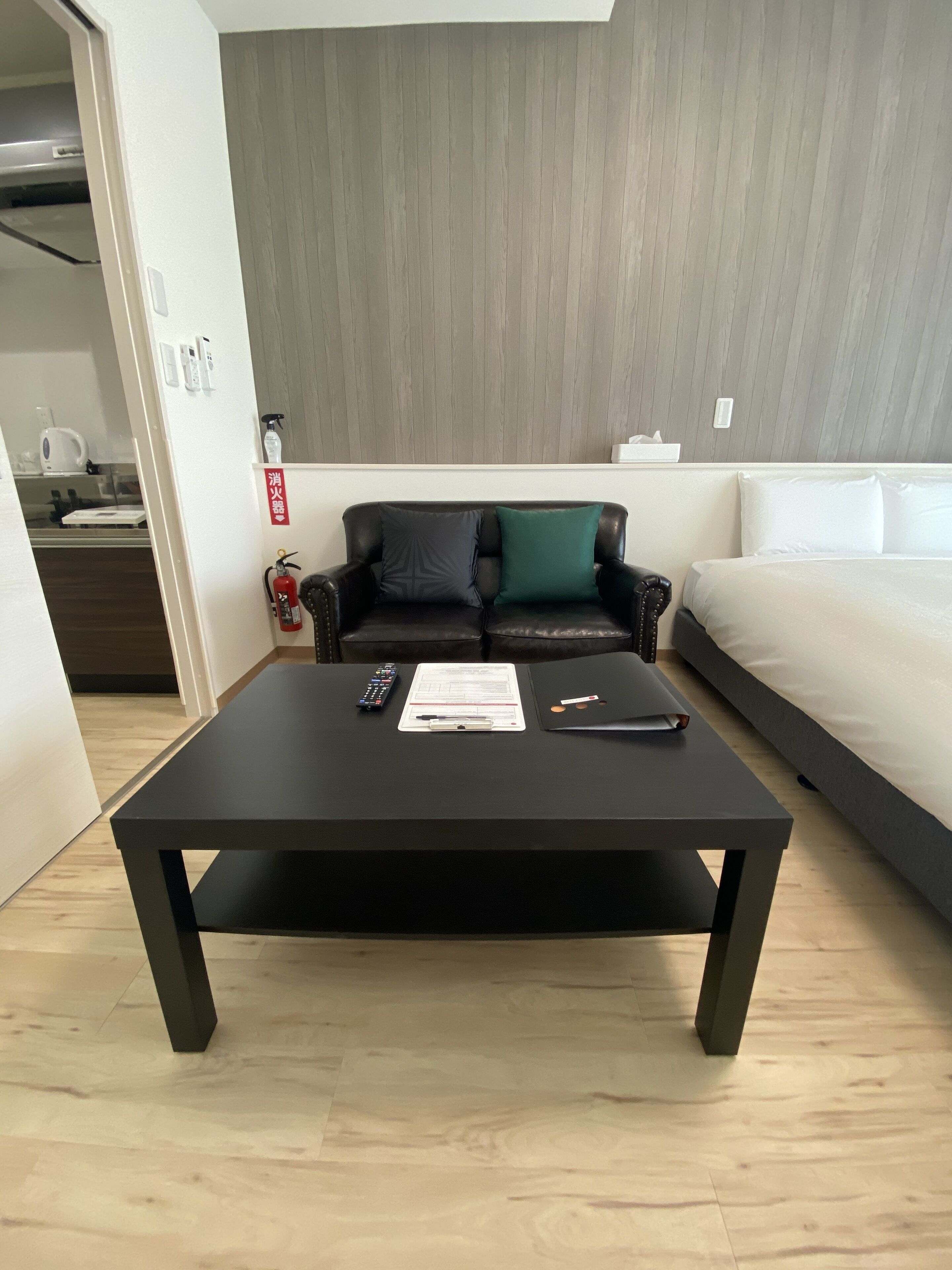 Un salon moderne avec une table basse noire, un canapé agrémenté de coussins foncés et verts, un lit blanc et un coin cuisine visible à travers une porte ouverte.