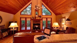 Interior acogedor de una cabaña con chimenea, techo de madera y ventanas amplias con vistas al exterior.