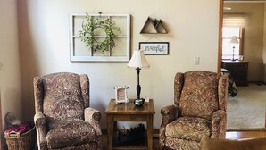 Un salón acogedor con dos sillones estampados, una mesita de madera con una lámpara y un adorno en la pared.