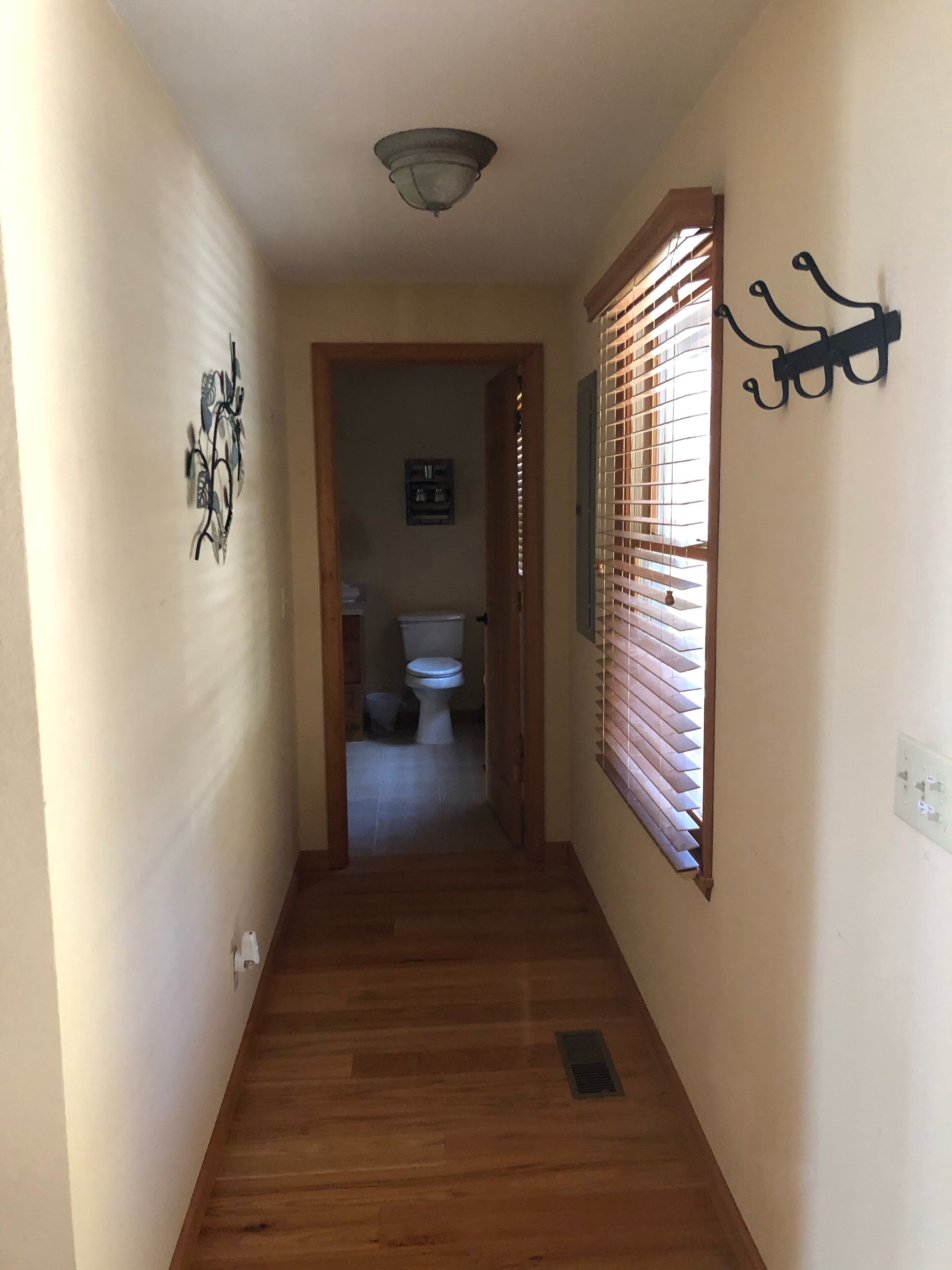 A hallway with wooden flooring, a ceiling light, a window with blinds, and a wall-mounted light fixture.