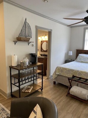 Een slaapkamer met een bed, nachtkastje, bedtafel met lamp, stoel, houten vloer en plafondventilator.