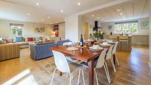 Un salón espacioso con una mesa de comedor de madera, sillas blancas y un sofá. Se ve una cocina con un ventanal grande y una estufa empotrada al fondo.