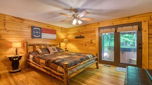 Une chambre de style cabane en bois, avec un lit, deux lampes de chevet, un ventilateur de plafond et une vue sur l’extérieur grâce à une porte-fenêtre coulissante.
