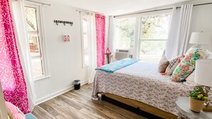 Ein Schlafzimmer mit einem Bett, einem kleinen Tisch mit Lampe, einem Fenster mit Vorhängen und einem Mülleimer.