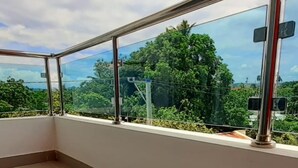 A balcony with a glass railing, a tiled floor, and a view of greenery and buildings. - Residencial Don Mero (La Vega)