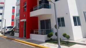 A modern multi-story building with a red and white facade, balconies, and a visible entrance. - Residencial Don Mero (La Vega)