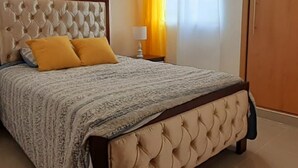 A bedroom with a tufted headboard, a bed with a patterned bedspread, two yellow pillows, and a nightstand with a lamp. - Residencial Don Mero (La Vega)