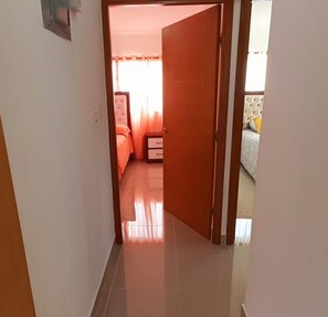 A narrow hallway leading to a bedroom with a bed, a nightstand, and a window with curtains. - Residencial Don Mero (La Vega)