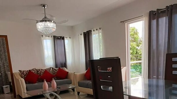 A modern living room with a glass dining table, a sofa with red cushions, a ceiling fan, and a view of the outdoors through large windows. - Residencial Don Mero (La Vega)