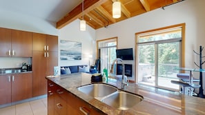 A modern kitchen with granite countertops, stainless steel sinks, wooden cabinets, a large window, and a flat-screen TV. - Suncatcher Penthouse in Sun Peaks Village (Sun Peaks)