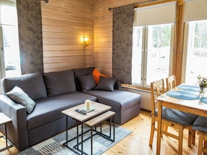 Een gezellige kamer met een grijze bank, een klein tafeltje met een afstandsbediening, en een eethoek met houten stoelen.