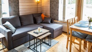 Ein gemütliches Zimmer mit grauem Sofa, einem kleinen Tisch mit Fernbedienung, einem Holzessttisch mit Stühlen und einem Fenster mit Blick auf Bäume.
