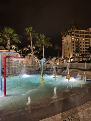 Ein Schwimmbecken bei Nacht mit mehreren Wasserspielen und Palmen im Hintergrund.