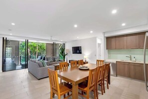 A modern kitchen with wooden cabinets, a dining table with chairs, a TV mounted on the wall, and a sliding glass door leading to a balcony.