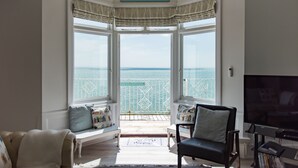 Ein Wohnzimmer mit Erkerfenster und Meerblick, einem Fernseher und einer Couch mit Kissen.